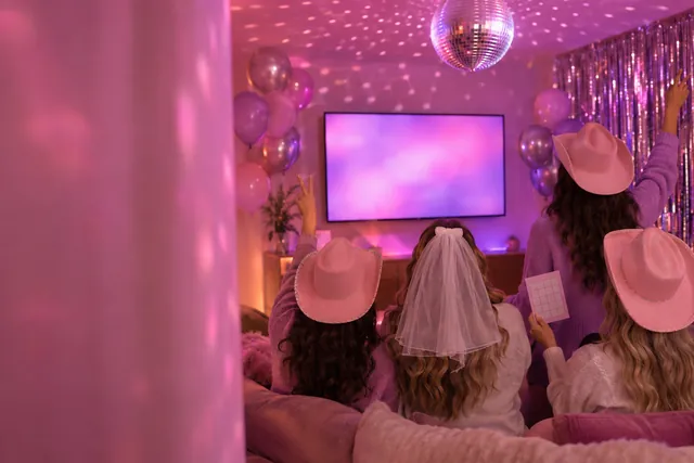 Friends in pink cowboy hats on a couch in a rental house, the bride in a veil, a disco ball and a TV glowing pink.
